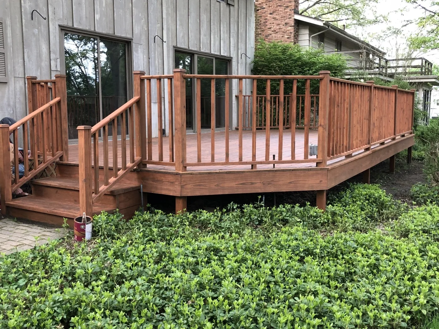 A wooden deck with railings and steps attached to the back of a house, surrounded by green landscaping.