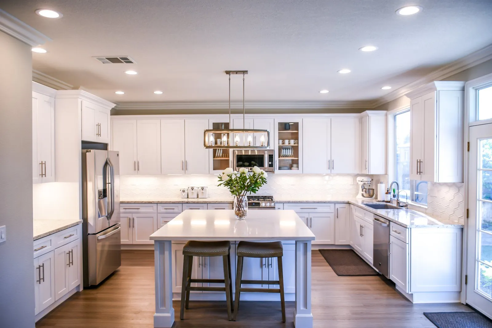 Modern white kitchen with a center island, two stools, stainless steel appliances, and wood floors.