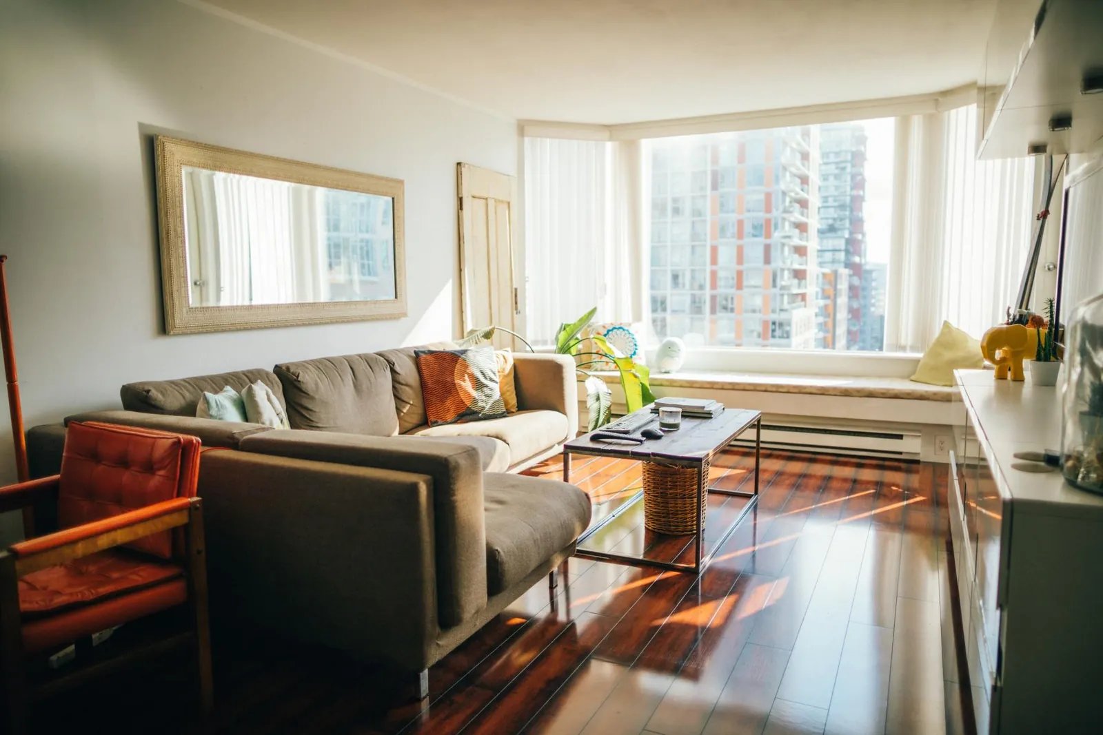 A sunlit living room with a gray sectional sofa, a coffee table, and a large mirror on a white wall above wooden floors.