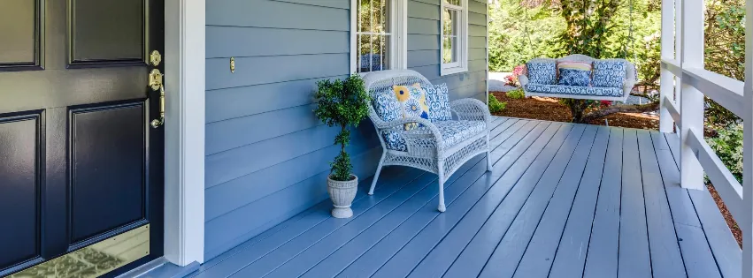 A light blue porch features a white wicker chair with floral cushions, a small potted plant, and a dark front door.