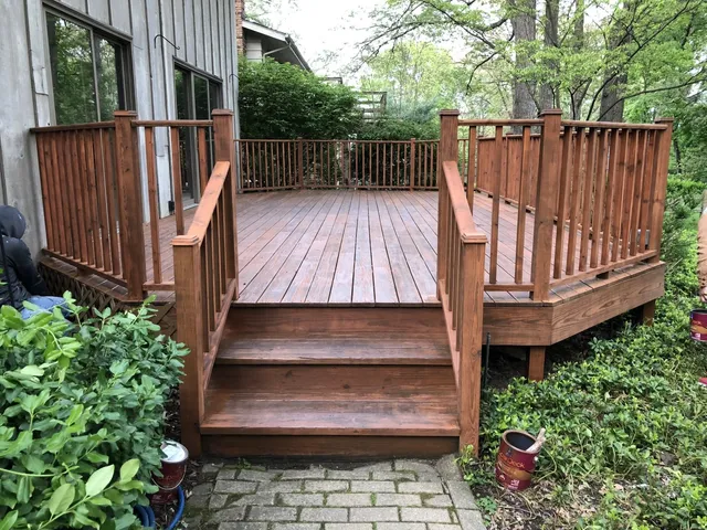 A newly stained brown wooden deck with stairs and railings attached to the back of a house, surrounded by greenery.