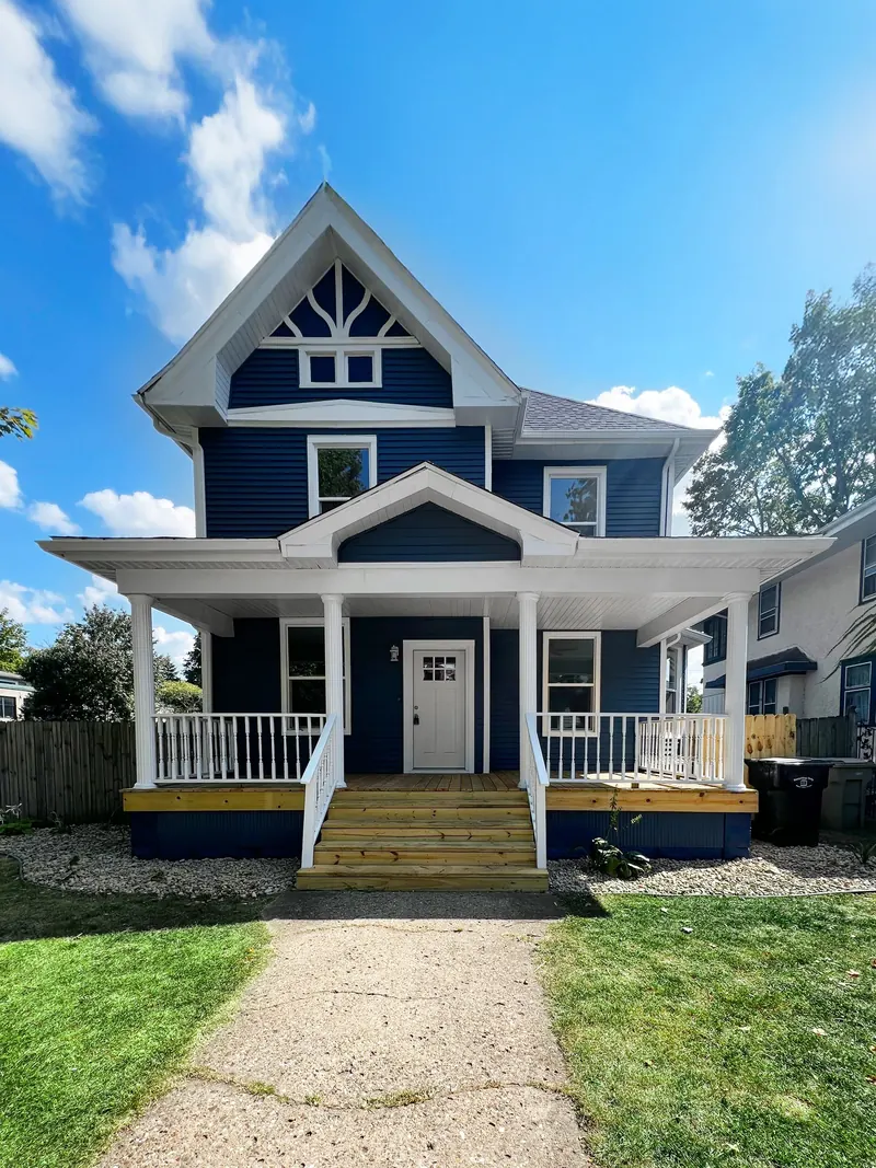 A two-story, dark blue house with white trim, a covered front porch, and wooden stairs, set under a sunny blue sky.
