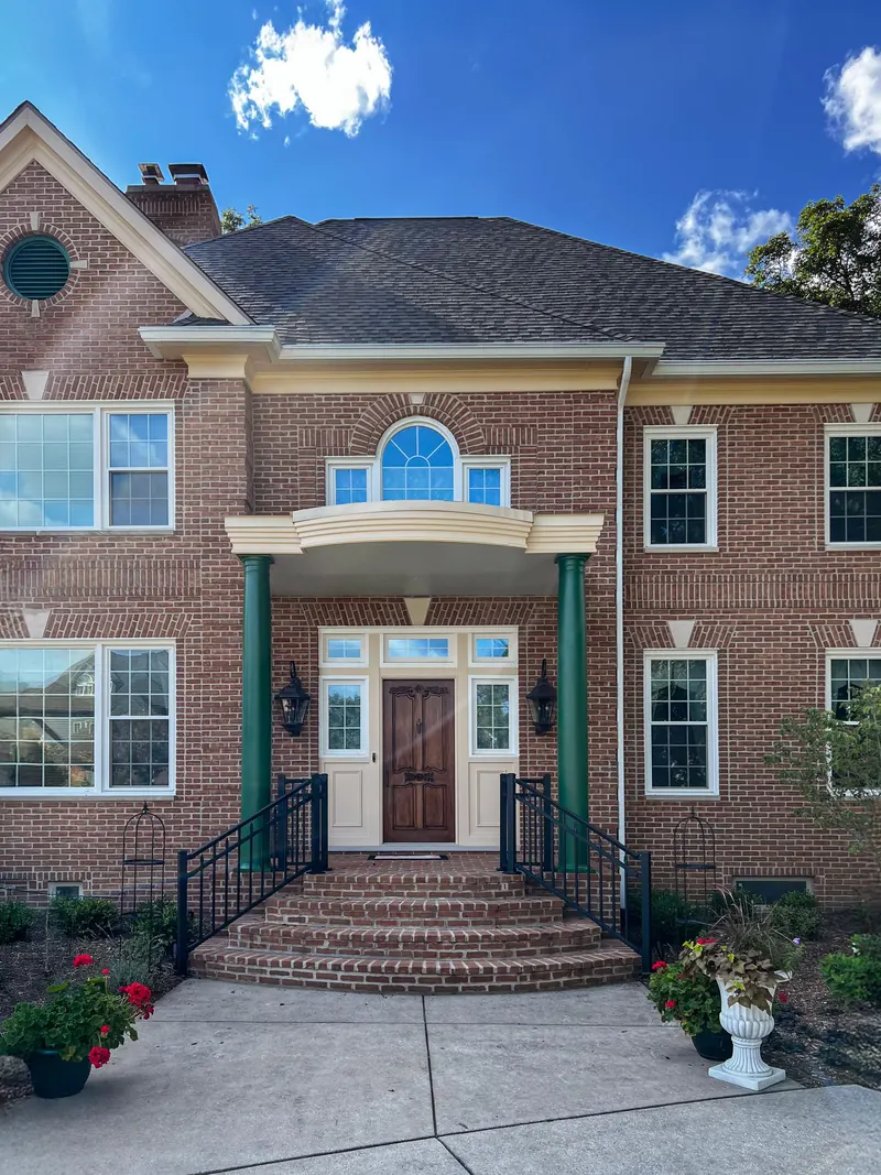 A two-story brick home with a covered front porch featuring two green columns and a wooden front door under a clear sky.