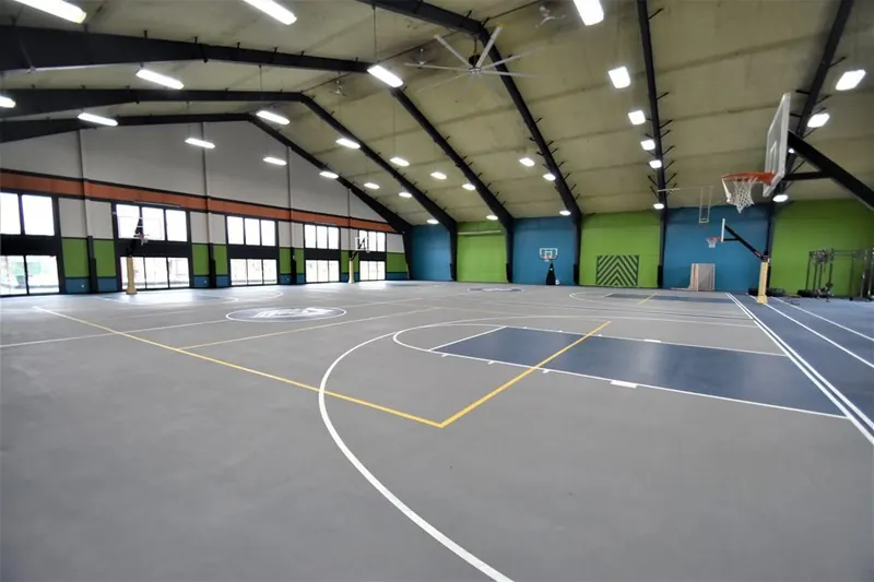 An indoor basketball court with grey flooring, yellow lines, basketball hoops, and light green and blue accent walls.