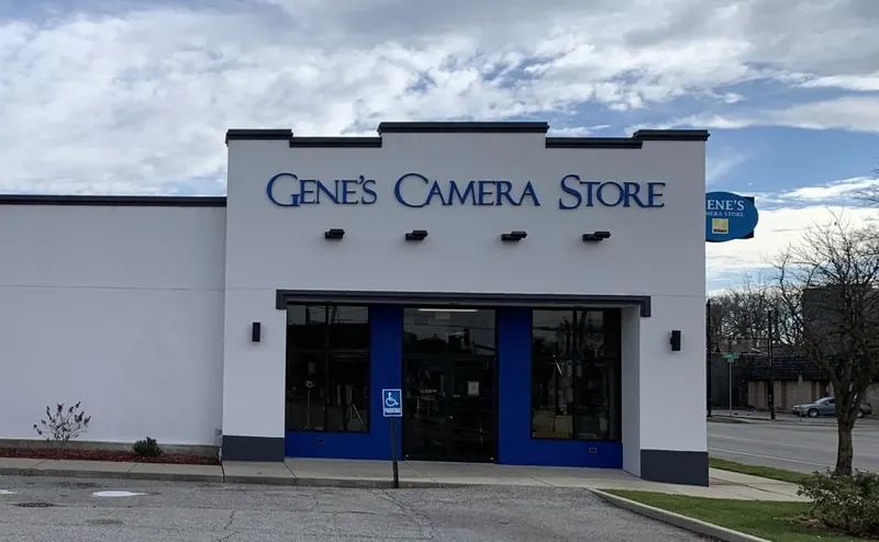 A white, single-story Gene's Camera Store building with a blue trim and entrance under a cloudy sky.