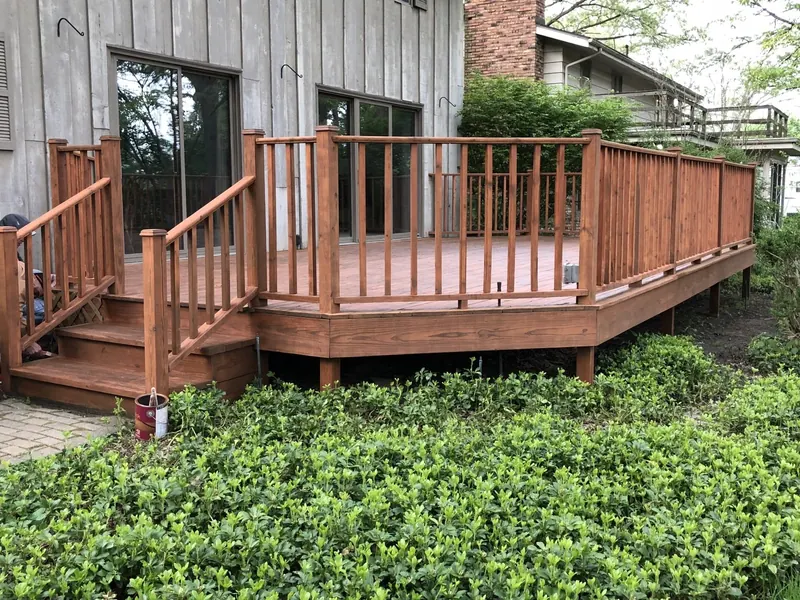 A stained wooden deck with railings and stairs sits outside the back of a house above a green garden.