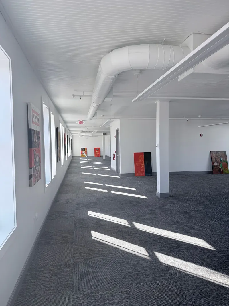 A long, sunlit hallway in an office or gallery space with grey patterned carpet, white walls, and an exposed ceiling pipe.