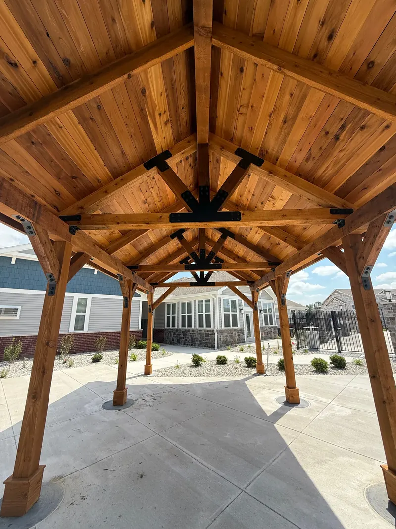 A wooden pavilion frame with exposed rafters and black metal hardware, standing over a concrete patio outside a house.
