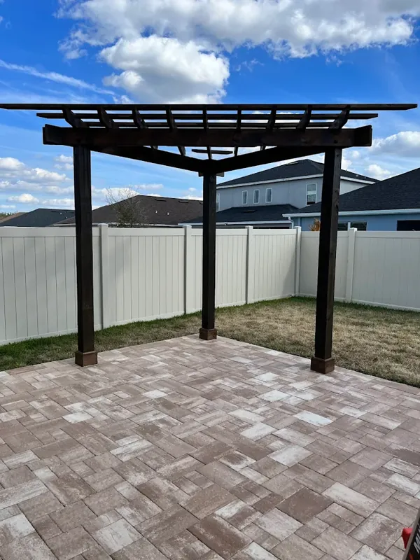 Dark-stained wood pergola over a paver patio with a white vinyl privacy fence.