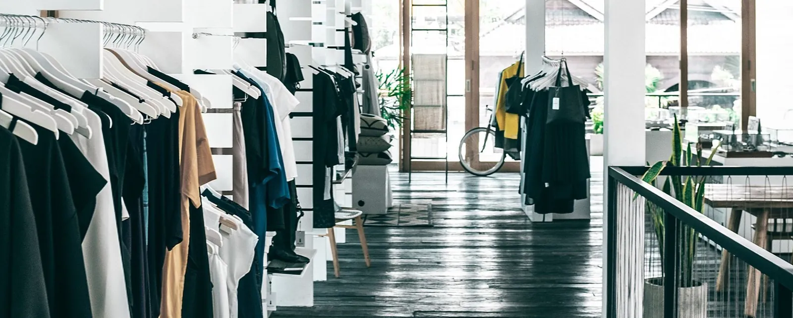 Minimalist clothing store interior with racks of neutral-toned garments, wooden flooring, and large glass windows.
