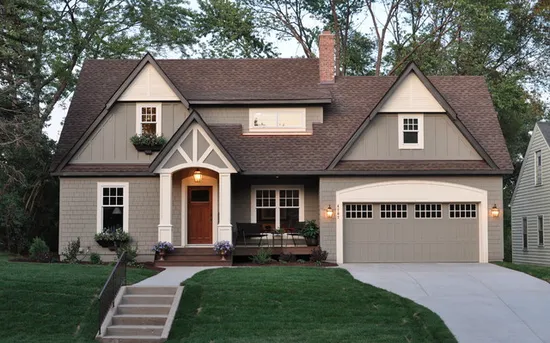 A two-story taupe house with a front porch, a two-car garage, and a brown shingled roof, surrounded by trees and lawn.