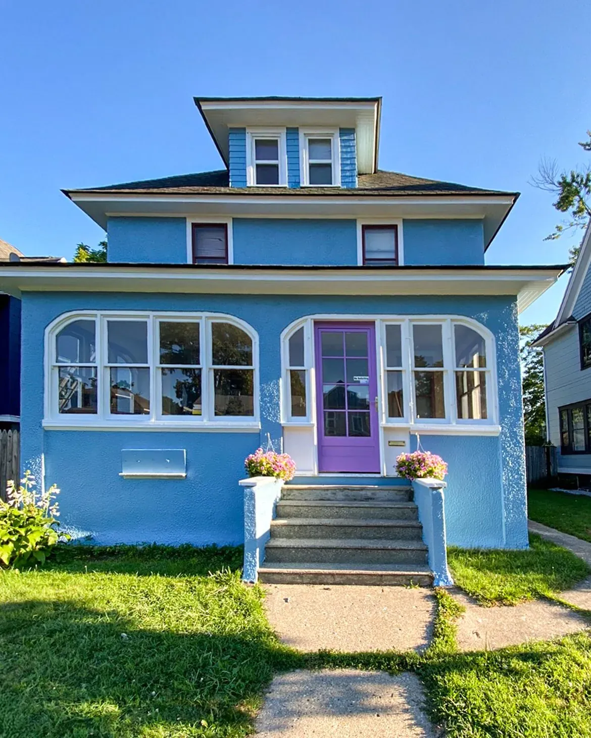 A blue house with a purple front door, white-trimmed windows, a central dormer, and concrete steps leading to the porch.