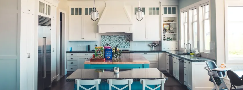 A bright kitchen with white cabinetry, a teal island, a matching dining table, and a stainless steel refrigerator.