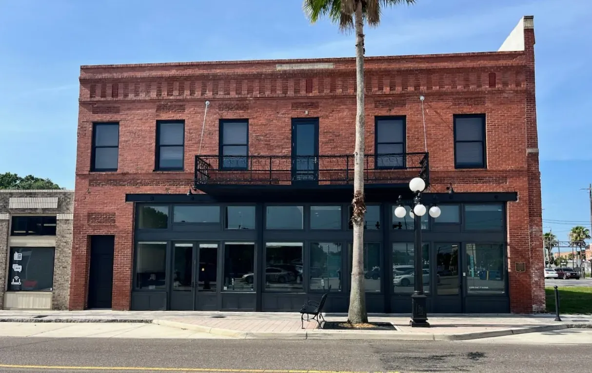 A two-story red brick building with a black storefront and iron balcony under a blue sky.