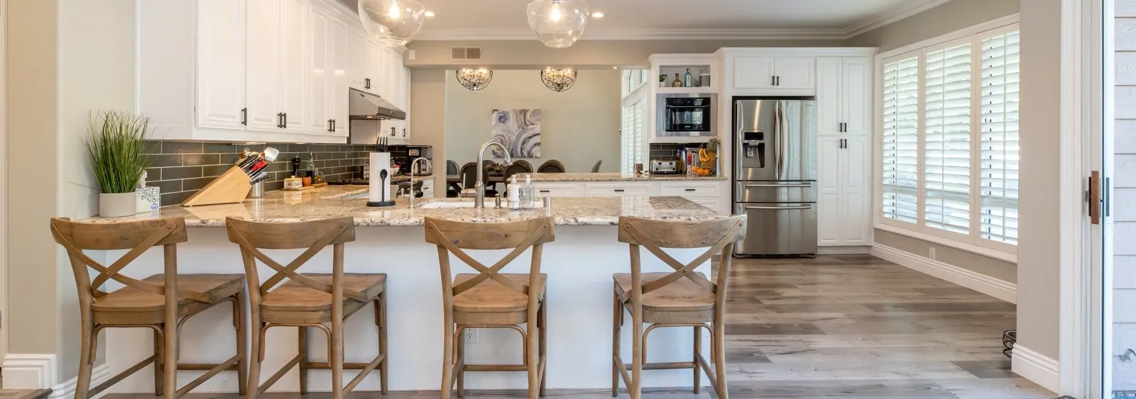 A bright kitchen with white cabinets, a stone island, four wooden stools, stainless steel appliances, and wood flooring.