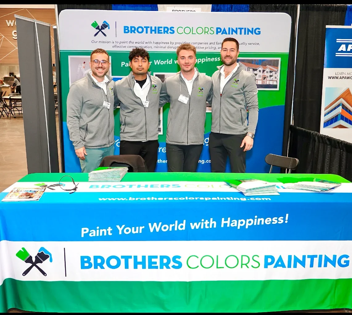 The Brothers Colors Painting team standing together at a trade show booth in matching gray quarter-zips, with a branded backdrop and table reading Paint Your World with Happiness.